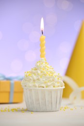 Photo of Burning candle in cupcake, sprinkles and party decor on white table against violet background with blurred lights