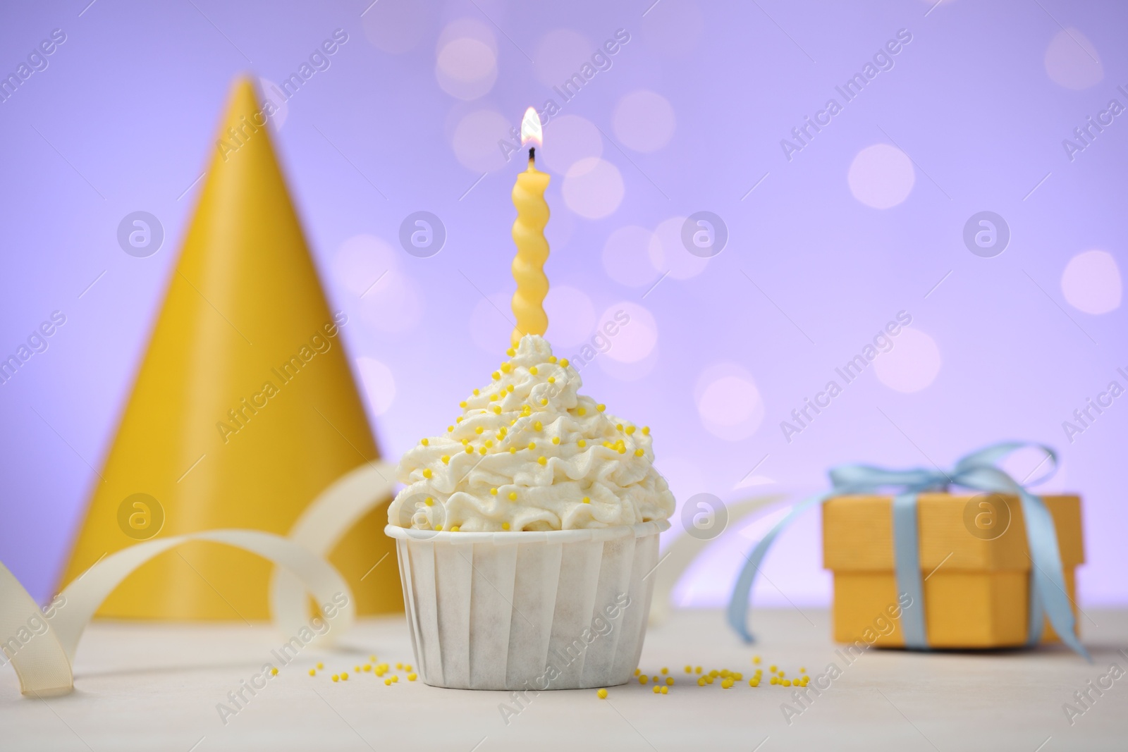 Burning candle in cupcake, sprinkles and party decor on white table against violet background with blurred lights Photo of Burning candle in cupcake, sprinkles and party decor on white table against violet background with blurred lights