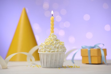 Photo of Burning candle in cupcake, sprinkles and party decor on white table against violet background with blurred lights