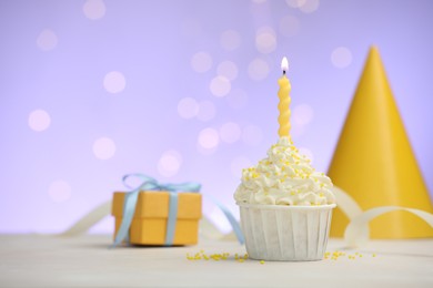 Photo of Burning candle in cupcake, sprinkles and party decor on white table against violet background with blurred lights, space for text