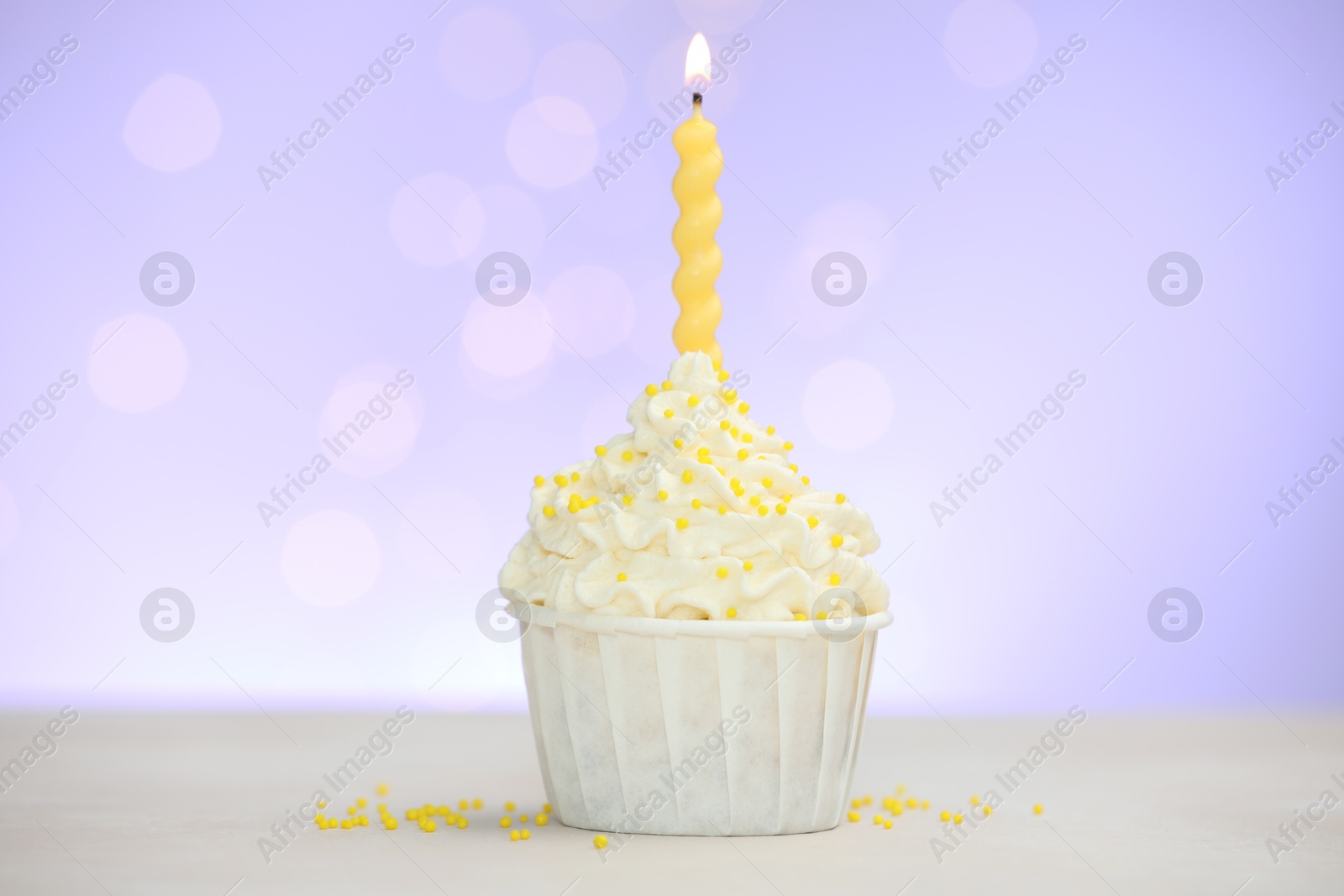 Burning candle in cupcake and sprinkles on white table against violet background with blurred lights Photo of Burning candle in cupcake and sprinkles on white table against violet background with blurred lights