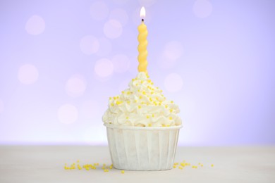Photo of Burning candle in cupcake and sprinkles on white table against violet background with blurred lights