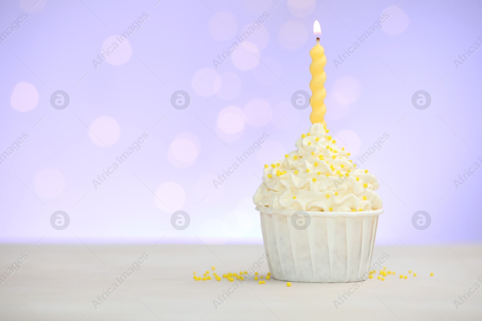 Photo of Burning candle in cupcake and sprinkles on white table against violet background with blurred lights, space for text