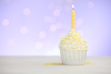 Photo of Burning candle in cupcake and sprinkles on white table against violet background with blurred lights, space for text