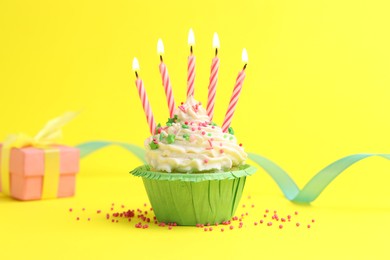 Photo of Burning candles in cupcake, sprinkles and party decor on yellow background