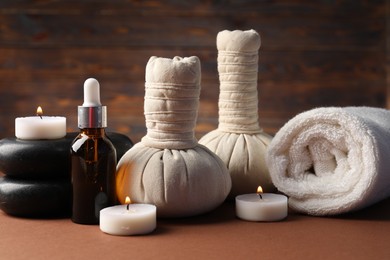 Photo of Massage bags, stones, towel, cosmetic product and burning candles on dark beige table, closeup. Spa composition