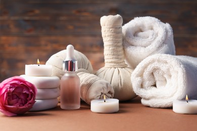 Photo of Massage bags, stones, towels, cosmetic product, peony flower and burning candles on dark beige table. Spa composition