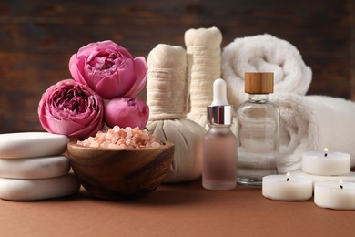 Photo of Massage bags, stones, towels, cosmetic products, burning candles and peony flowers on dark beige table. Spa composition