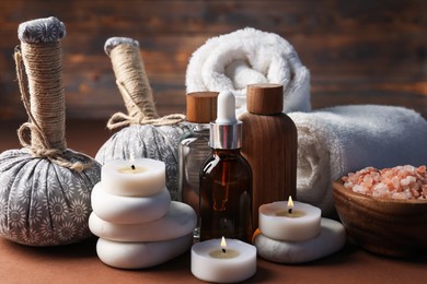 Photo of Massage bags, stones, towels, cosmetic products and burning candles on dark beige table, closeup. Spa composition