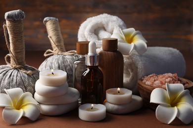 Photo of Massage bags, stones, towels, cosmetic products, plumeria flowers and burning candles on dark beige table, closeup. Spa composition