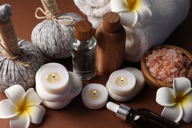 Photo of Massage bags, stones, towels, cosmetic products, plumeria flowers and burning candles on dark beige table, closeup. Spa composition