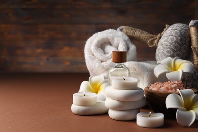 Photo of Spa composition. Massage bags, stones, towels, cosmetic products, plumeria flowers and burning candles on dark beige table, closeup. Space for text