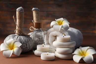 Photo of Massage bags, stones, towel, cosmetic product, plumeria flowers and burning candles on dark beige table, closeup. Spa composition