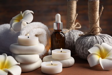Photo of Massage bags, stones, towel, cosmetic product, plumeria flowers and burning candles on dark beige table, closeup. Spa composition