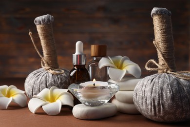 Photo of Massage bags, stones, cosmetic products, burning candle and plumeria flowers on dark beige table, closeup. Spa composition