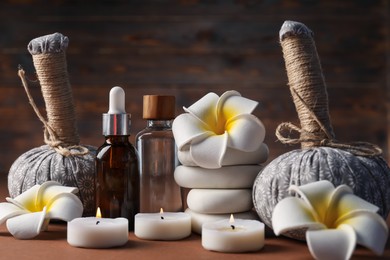 Photo of Massage bags, stones, cosmetic products, burning candles and plumeria flowers on dark beige table, closeup. Spa composition