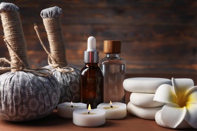 Photo of Massage bags, stones, cosmetic products, burning candles and plumeria flower on dark beige table, closeup. Spa composition