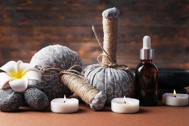 Photo of Massage bags, stones, cosmetic product, burning candles and plumeria flower on dark beige table, closeup. Spa composition