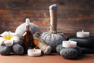 Photo of Massage bags, stones, cosmetic product, burning candles and plumeria flower on dark beige table, closeup. Spa composition