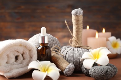 Photo of Massage bags, stones, cosmetic product, towel burning candles and plumeria flowers on dark beige table, closeup. Spa composition