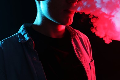 Photo of Man using electronic smoking device in neon lights on black background, closeup