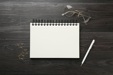 Photo of Open blank notebook, glasses, paperclips and pen on black wooden table, flat lay