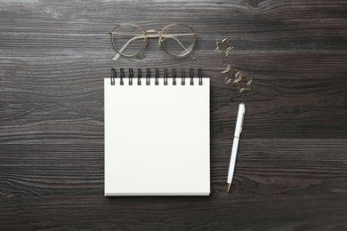 Photo of Open blank notebook, glasses, paperclips and pen on black wooden table, flat lay