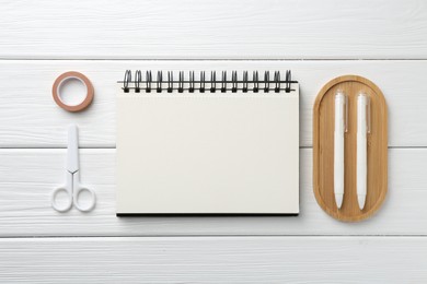 Photo of Open blank notebook, scissors, tape and pens on white wooden table, flat lay
