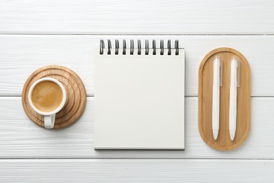 Photo of Open blank notebook, coffee and pens on white wooden table, flat lay
