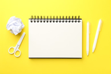 Photo of Open blank notebook, crumpled paper, scissors and pens on yellow background, flat lay