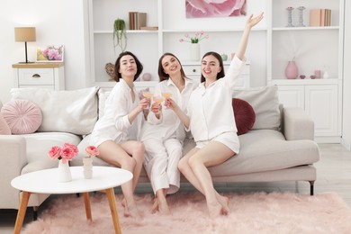 Photo of Pajama party. Happy friends clinking glasses on sofa at home