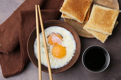 Photo of Half-boiled eggs in bowl, chopsticks, soy sauce and toasted bread on brown table, flat lay