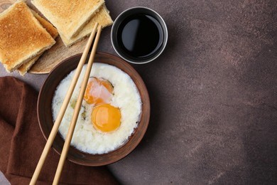 Photo of Half-boiled eggs in bowl, chopsticks, soy sauce and toasted bread on brown table, flat lay. Space for text