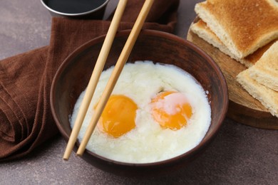 Photo of Half-boiled eggs in bowl, chopsticks, soy sauce and toasted bread on brown table, closeup
