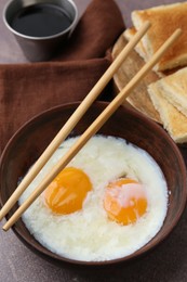 Photo of Half-boiled eggs in bowl, chopsticks, soy sauce and toasted bread on brown table, closeup