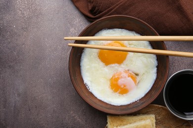 Photo of Half-boiled eggs in bowl, chopsticks, soy sauce and toasted bread on brown table, flat lay. Space for text