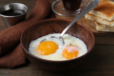 Photo of Half-boiled eggs in bowl, soy sauce and toasted bread on wooden table, closeup