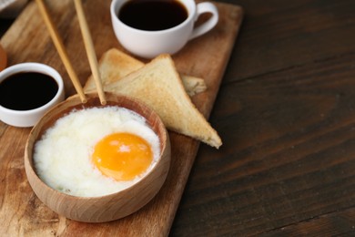 Photo of Half-boiled egg in bowl, soy sauce, chopsticks, coffee and toasted bread on wooden table, closeup. Space for text