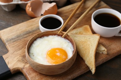 Photo of Half-boiled egg in bowl, soy sauce, chopsticks, coffee and toasted bread on wooden table, closeup