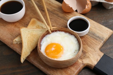 Photo of Half-boiled egg in bowl, soy sauce, chopsticks, coffee and toasted bread on wooden table, closeup