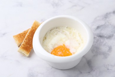 Photo of Half-boiled egg in bowl and toasted bread on white marble table, closeup