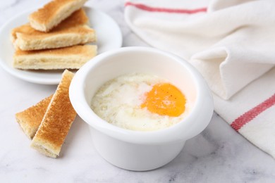 Photo of Half-boiled egg in bowl and toasted bread on white marble table, closeup