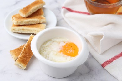 Photo of Half-boiled egg in bowl and toasted bread on white marble table, closeup
