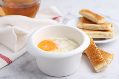 Photo of Half-boiled egg in bowl and toasted bread on white marble table, closeup