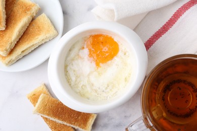 Photo of Half-boiled egg in bowl, tea and toasted bread on white marble table, flat lay