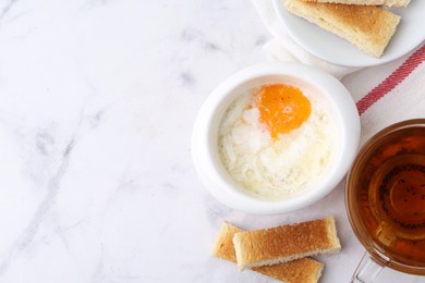 Photo of Half-boiled egg in bowl, tea and toasted bread on white marble table, flat lay