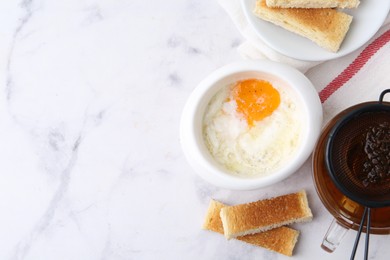 Photo of Half-boiled egg in bowl, tea and toasted bread on white marble table, flat lay