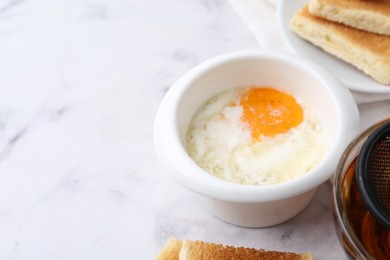 Photo of Half-boiled egg in bowl, tea and toasted bread on white marble table, closeup