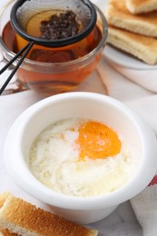 Photo of Half-boiled egg in bowl, tea and toasted bread on white marble table, closeup