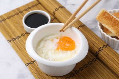 Photo of Half-boiled egg in bowl, soy sauce, chopsticks and toasted bread on white marble table, closeup